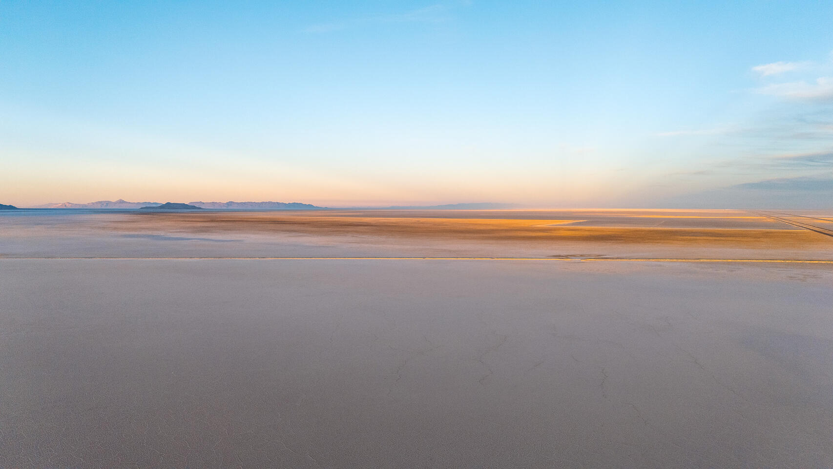 Aerial view of salt flats with a golden band of sunlight against pale blue sky.